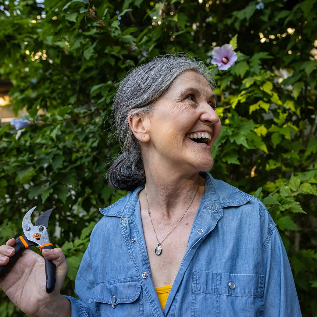 An older woman holding pruners out in her garden.