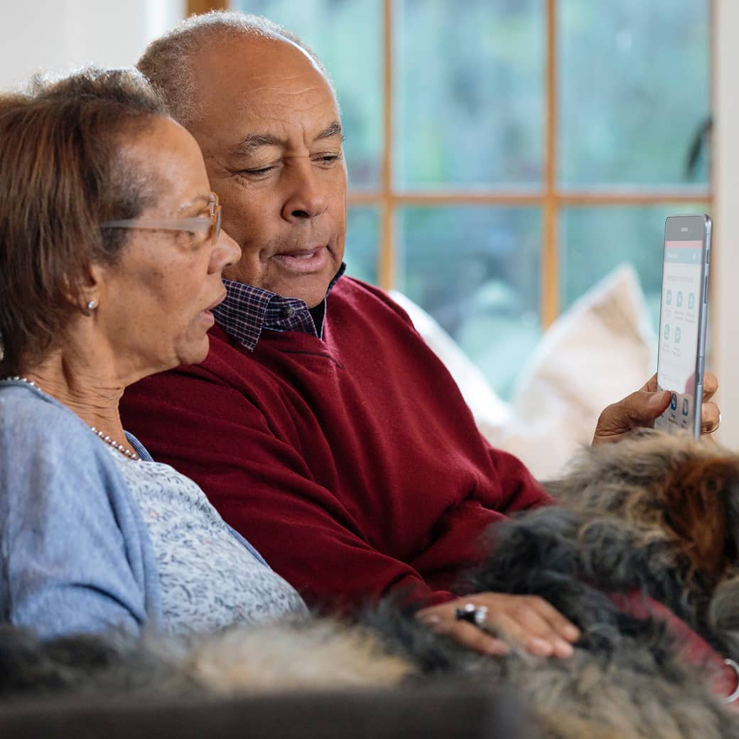 An older couple with their dog on the sofa, looking at a tablet screen together.