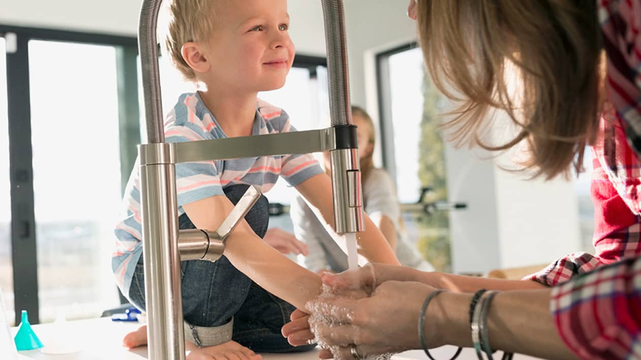 A small child smiles at their parent who is washing their hands in the kitchen sink.