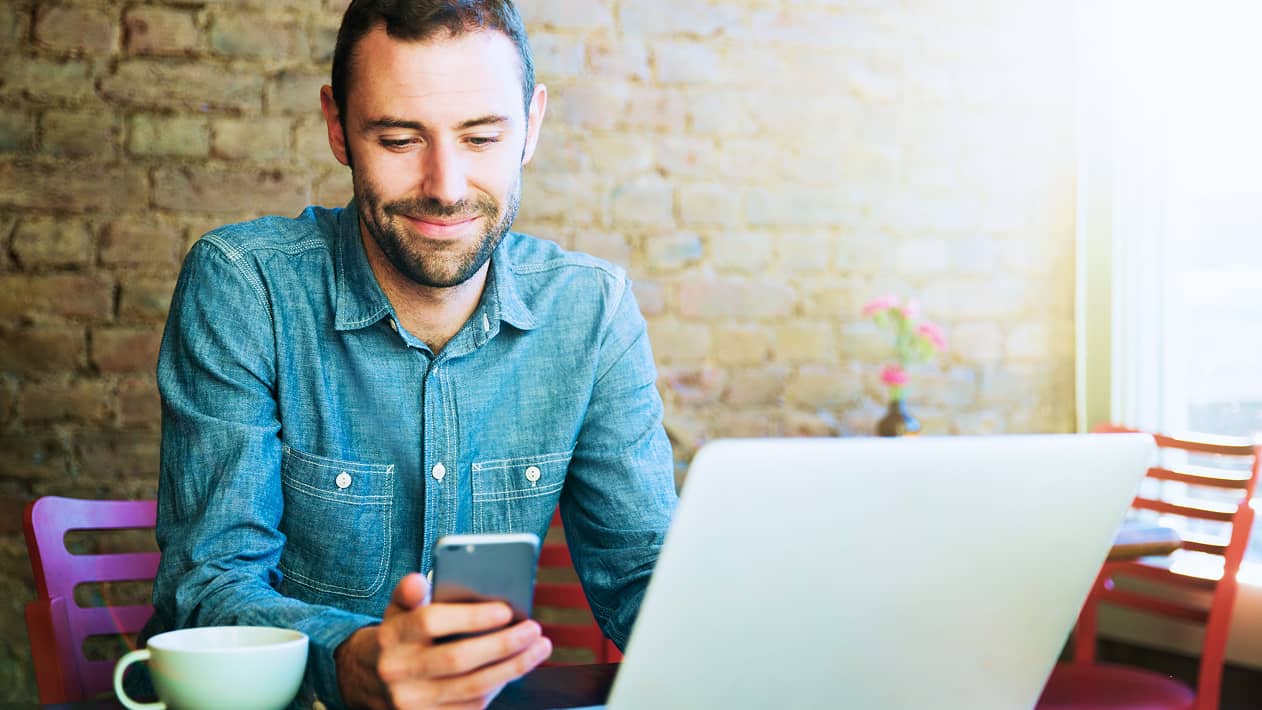 Someone looking at their cell phone screen with their laptop open as they sit in a coffeeshop