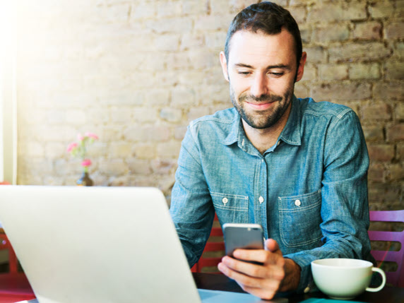 A man using a smartphone while working on a laptop in a café.