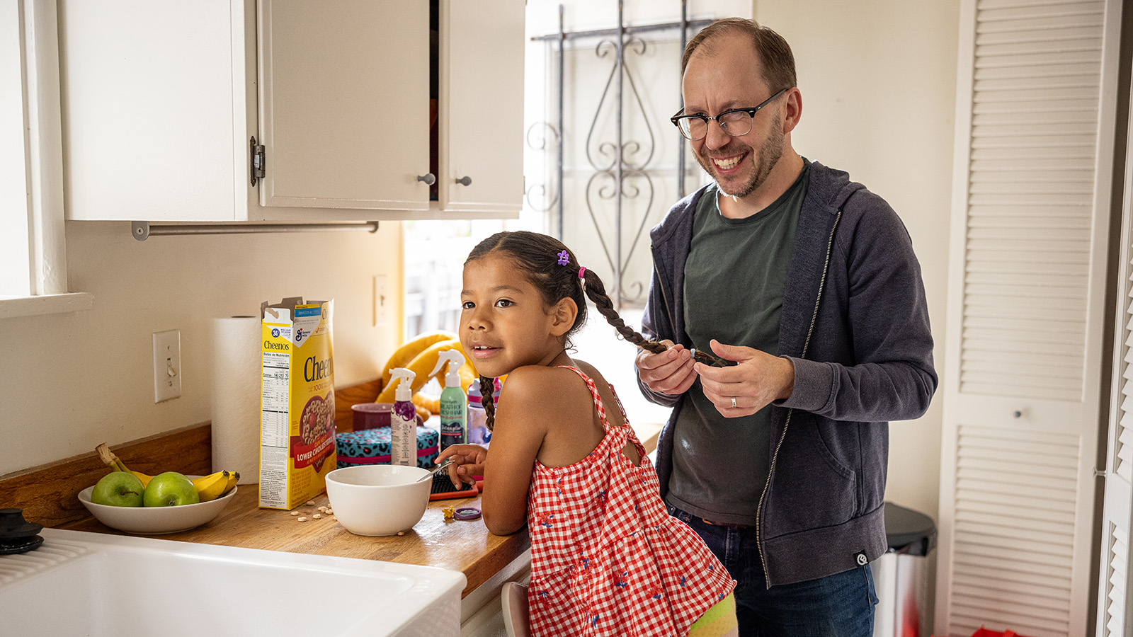Adult braiding child's hair and preparing food together in a kitchen.