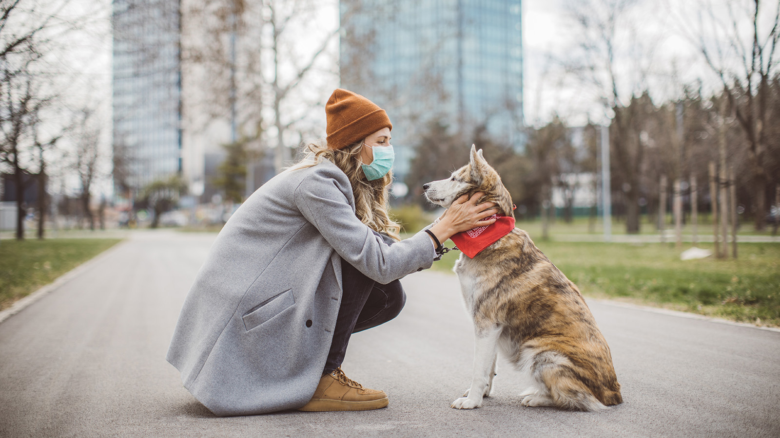 A woman in a gray coat kneels on a path, affectionately petting a dog wearing a red bandana.