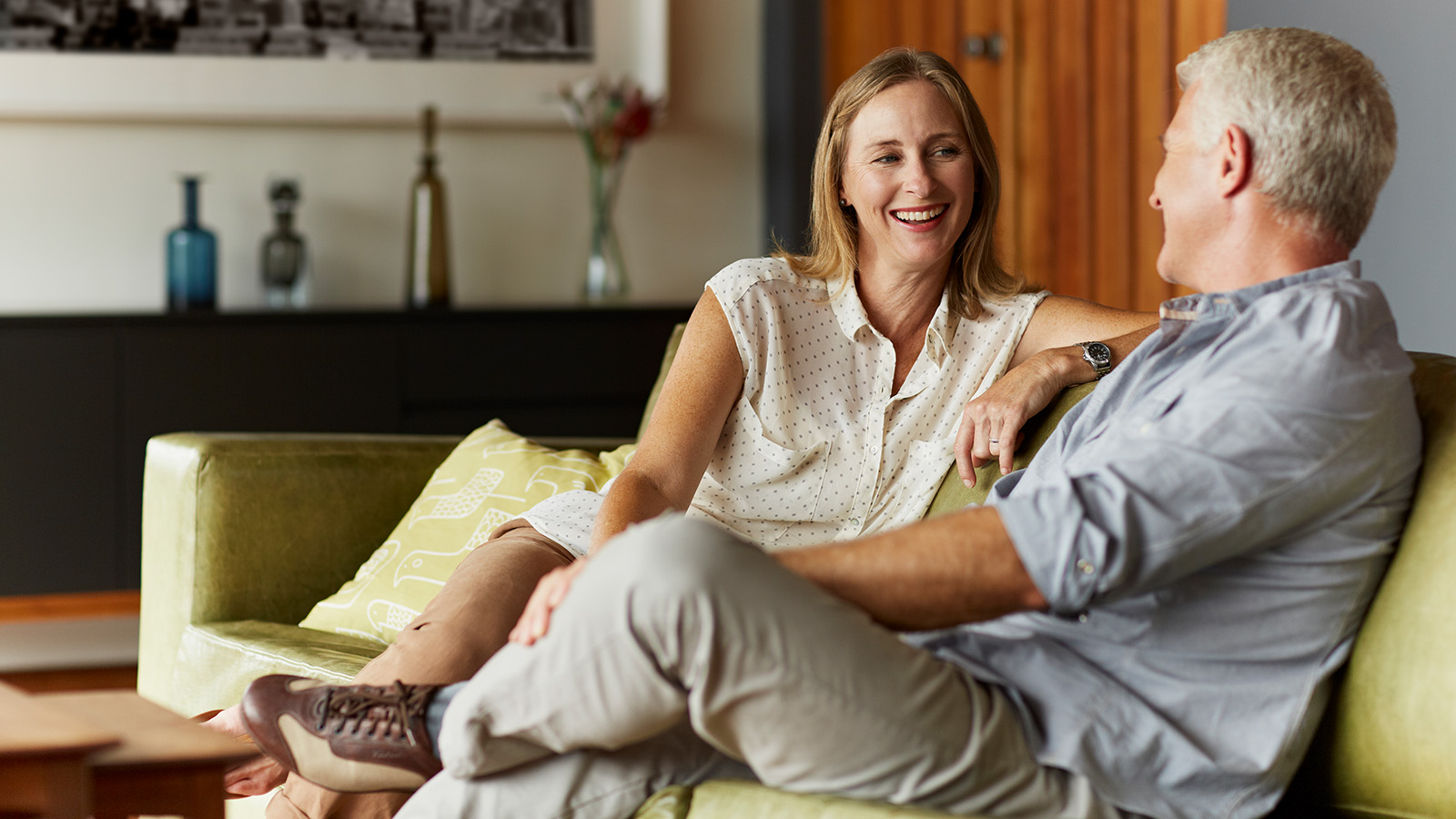 A couple sits on a green couch, engaged in conversation, with decorative items visible in the background.