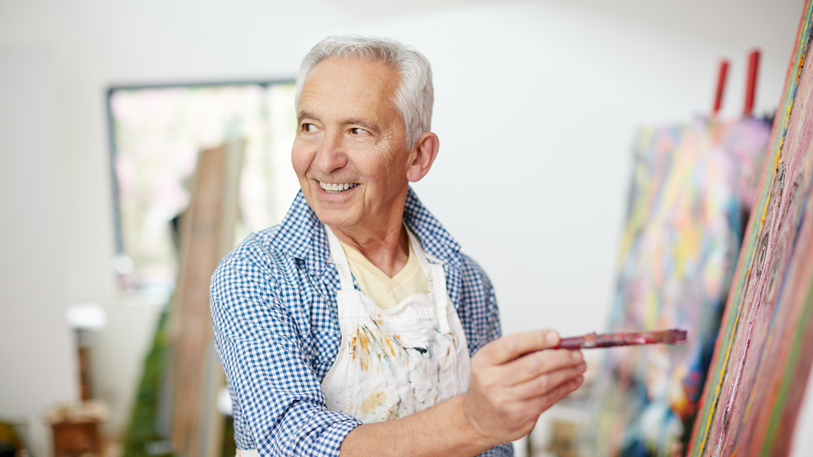 A man with gray hair holds a paintbrush, standing in front of a colorful canvas in an art studio.