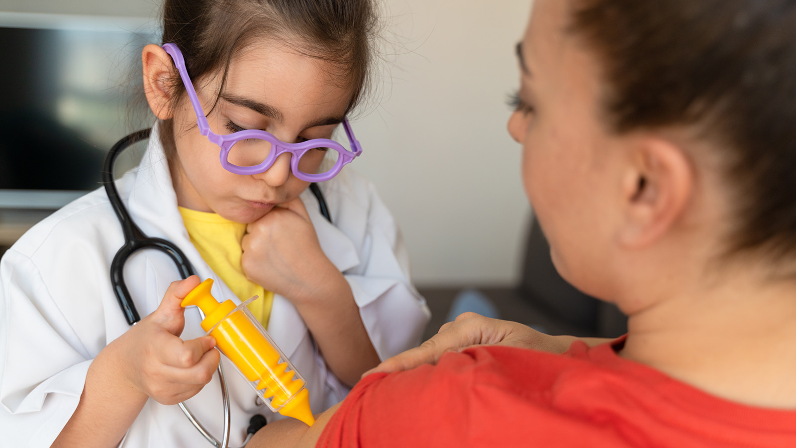 A child in a doctor’s coat pretends to give an injection with a toy syringe to an adult’s arm.