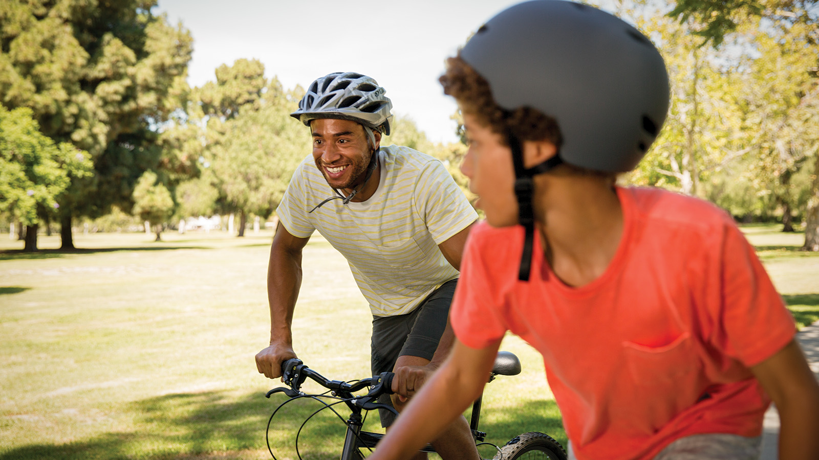 A father and son ride bicycles in a park, wearing helmets and enjoying a sunny day together.