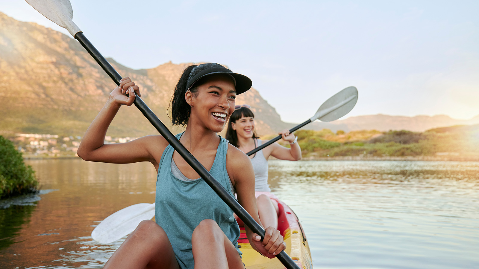 Two women paddle a kayak on a calm lake surrounded by mountains, enjoying a sunny day outdoors.