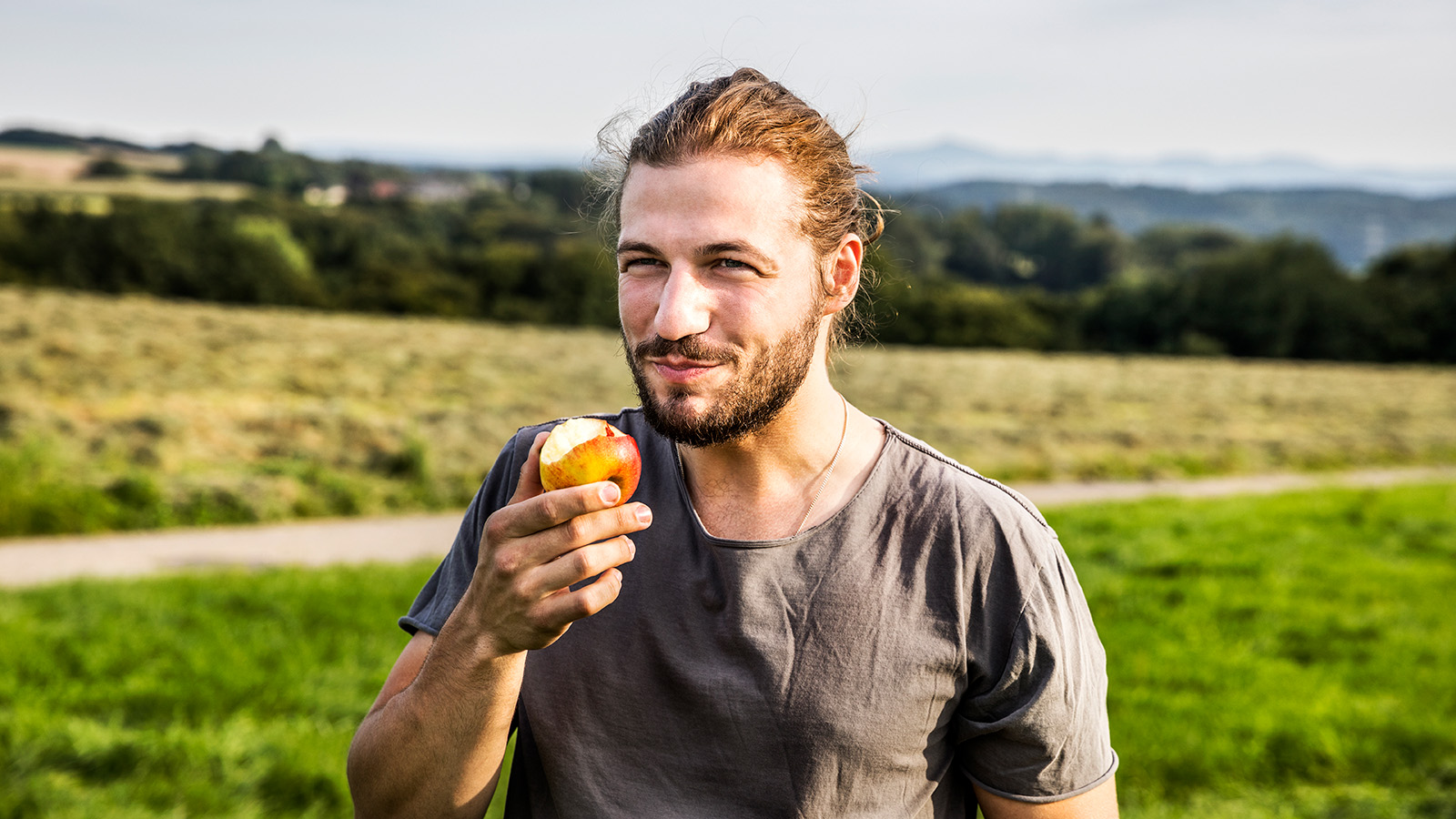 A person holds an apple, standing in a field with a scenic background of trees and hills.