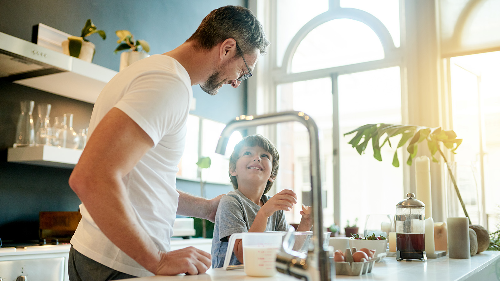 An adult and child prepare food together in a kitchen, with ingredients and utensils visible on the counter.