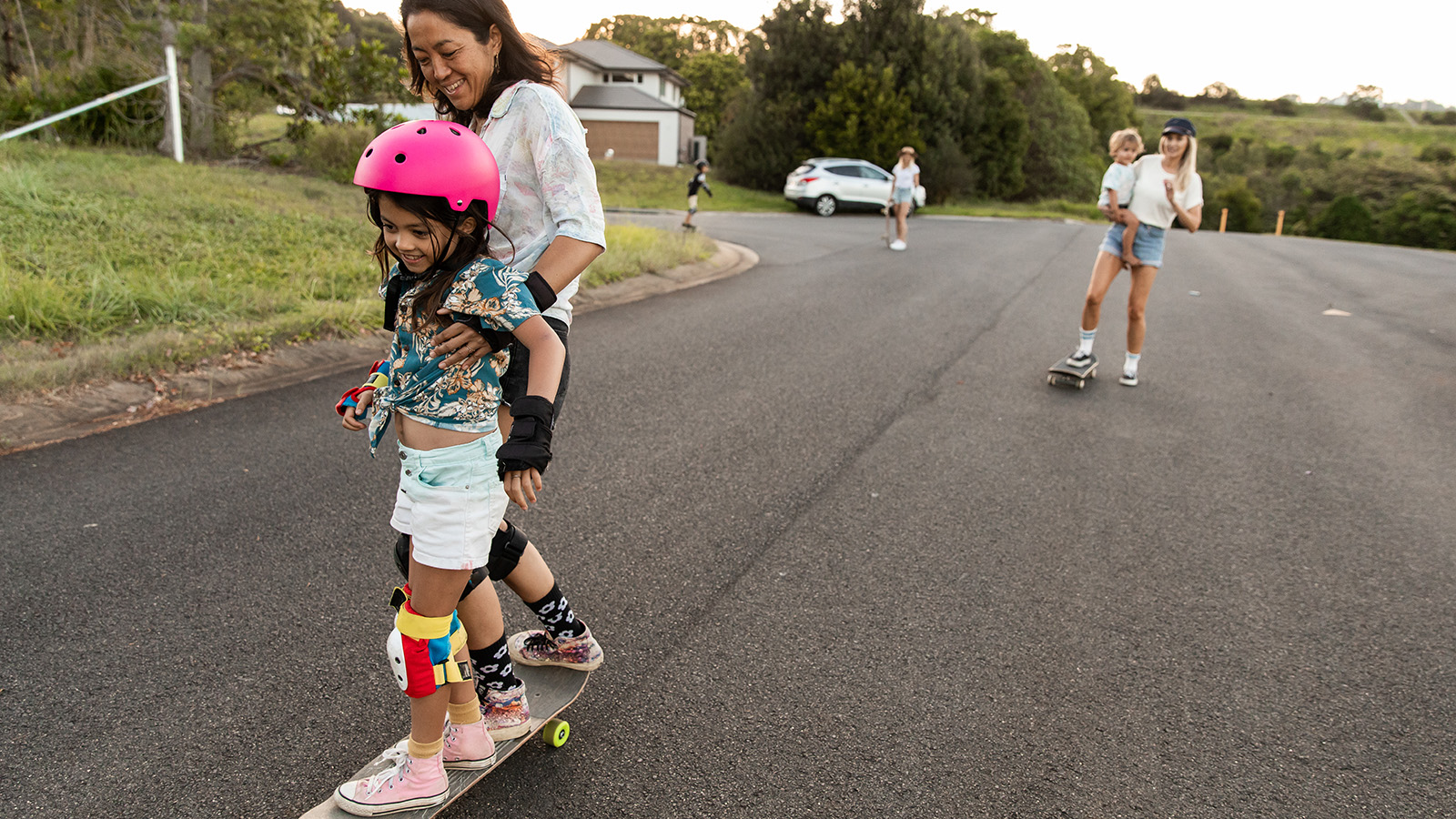 A child in a pink helmet rides a skateboard, supported by an adult, while others skate in a sunny neighborhood.