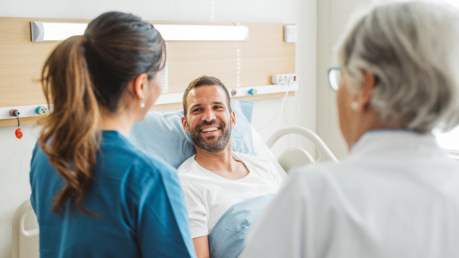 Two healthcare professionals converse with a patient in a hospital bed, discussing care in a bright, clinical environment.