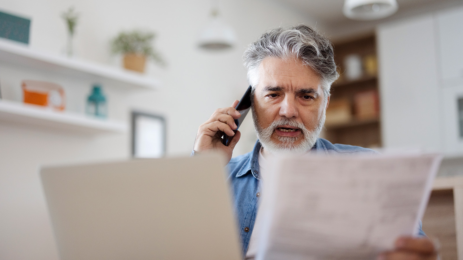 A man is on the phone while holding documents, seated at a table in a well-lit room.