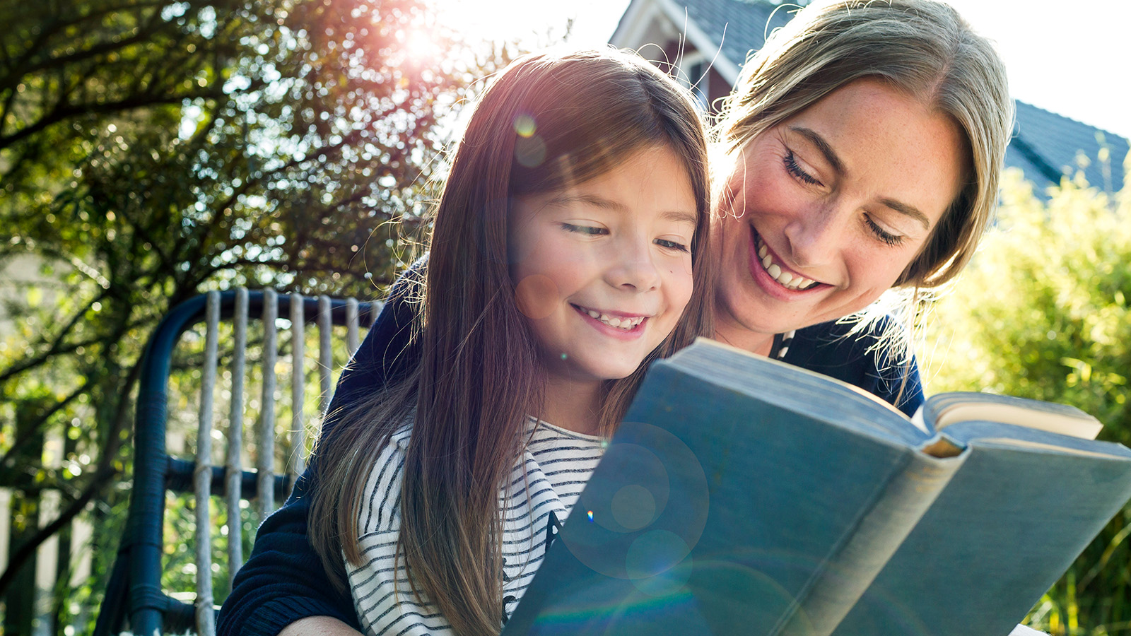 An adult reads a book with a child, both engaged and enjoying a sunny outdoor setting.