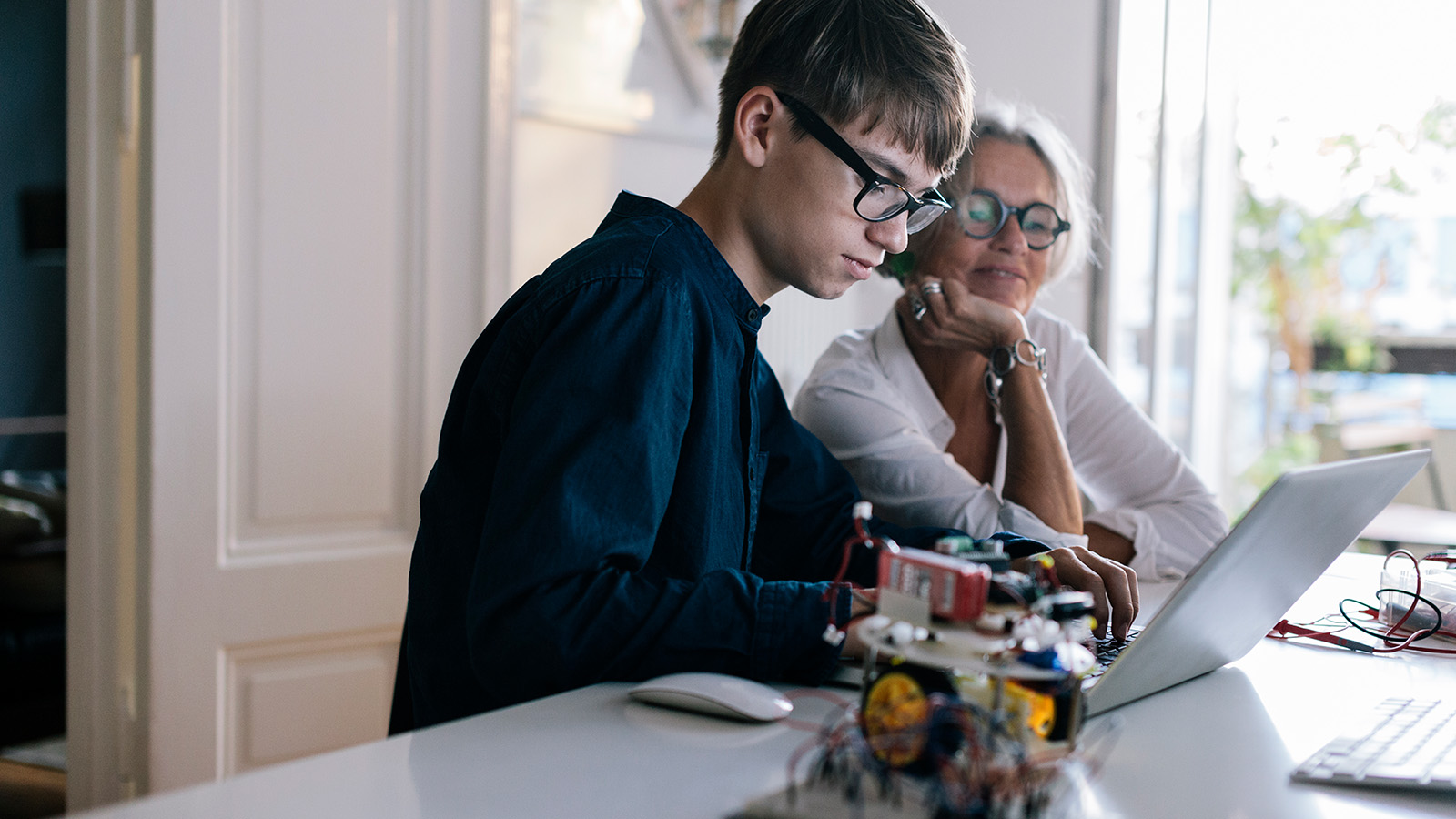A young person works on a laptop while an older woman observes, with electronic components scattered on the table.