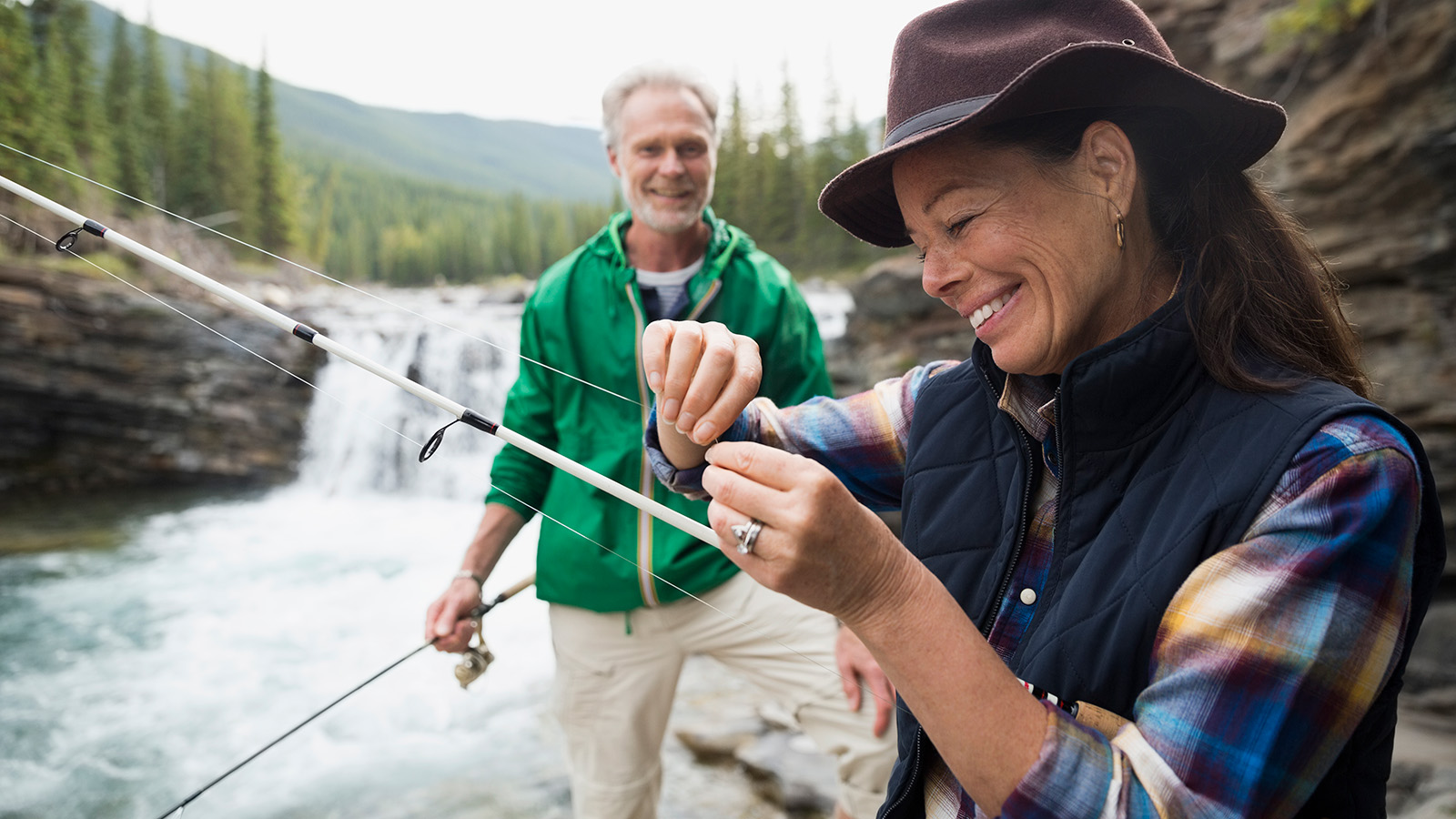 A woman adjusts fishing gear while a man stands nearby, surrounded by a natural landscape with a waterfall.