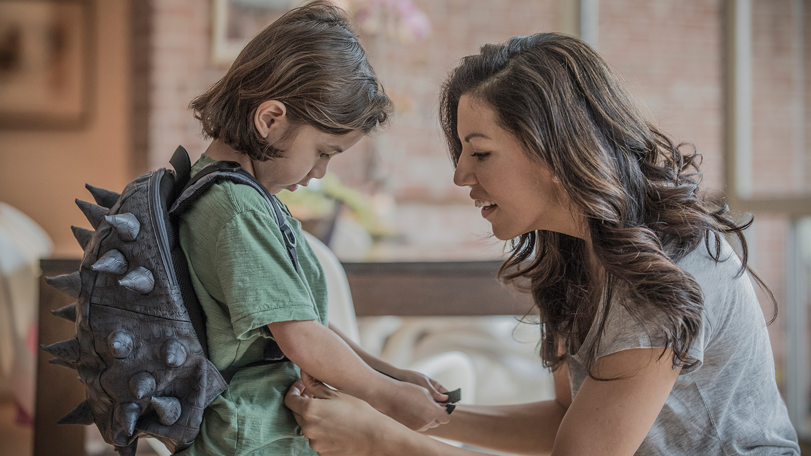 A woman assists a child wearing a spiked backpack in an indoor space with furniture and a brick wall.