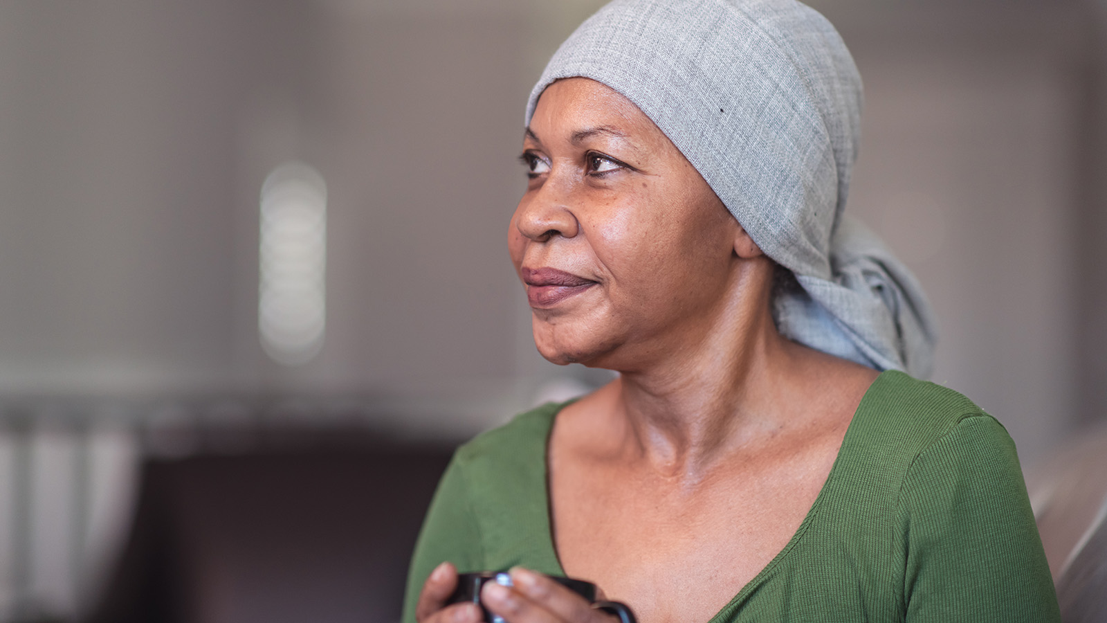 A woman wearing a headscarf sits thoughtfully, holding a small object in her hands, in a softly lit room.