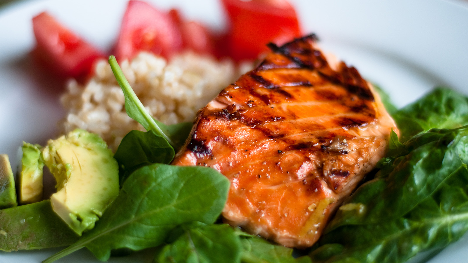 A plate features grilled salmon, brown rice, fresh spinach, and sliced tomatoes, showcasing a healthy meal.