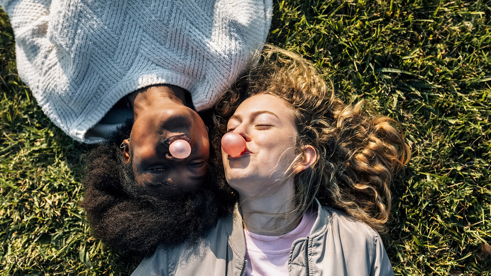 Two girls lie on the grass, smiling and enjoying each other’s company, with one blowing a bubble with gum.