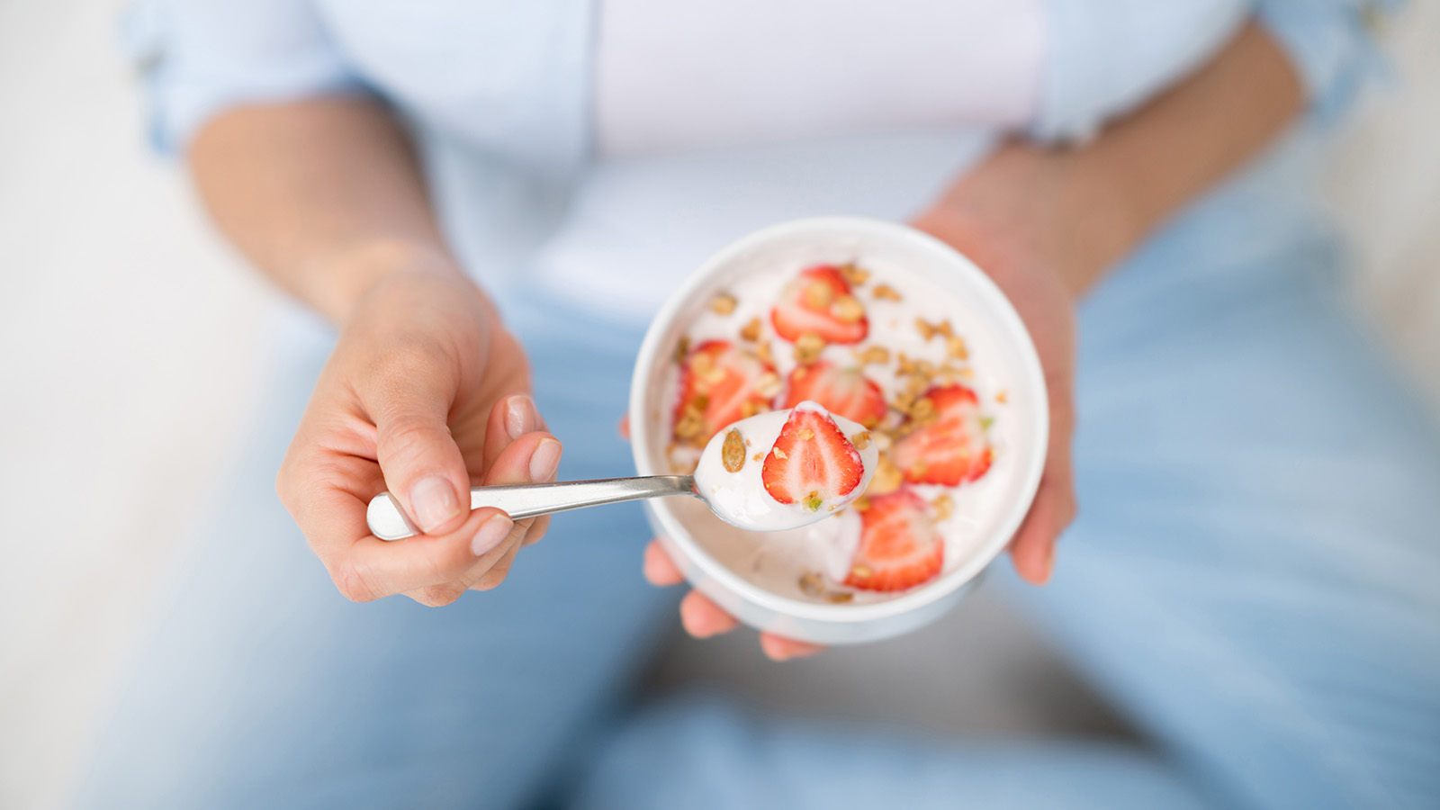 A person holds a bowl of yogurt topped with strawberries and granola, preparing to take a bite.