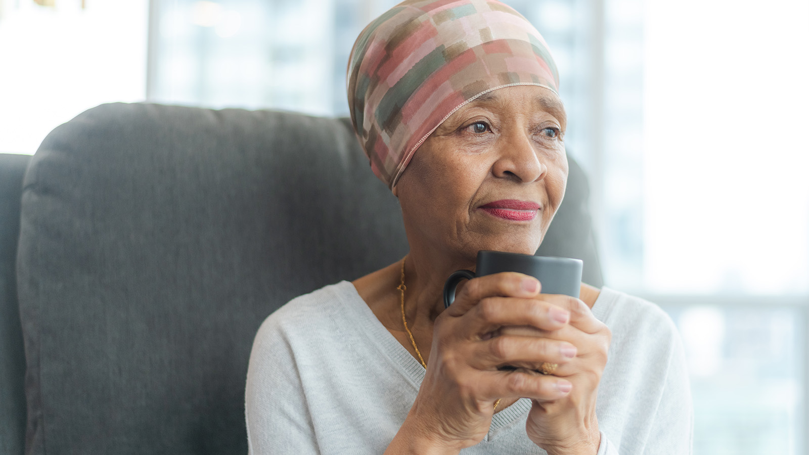 A person sits in a chair, holding a cup, looking thoughtfully while gazing out a window in a cozy setting.