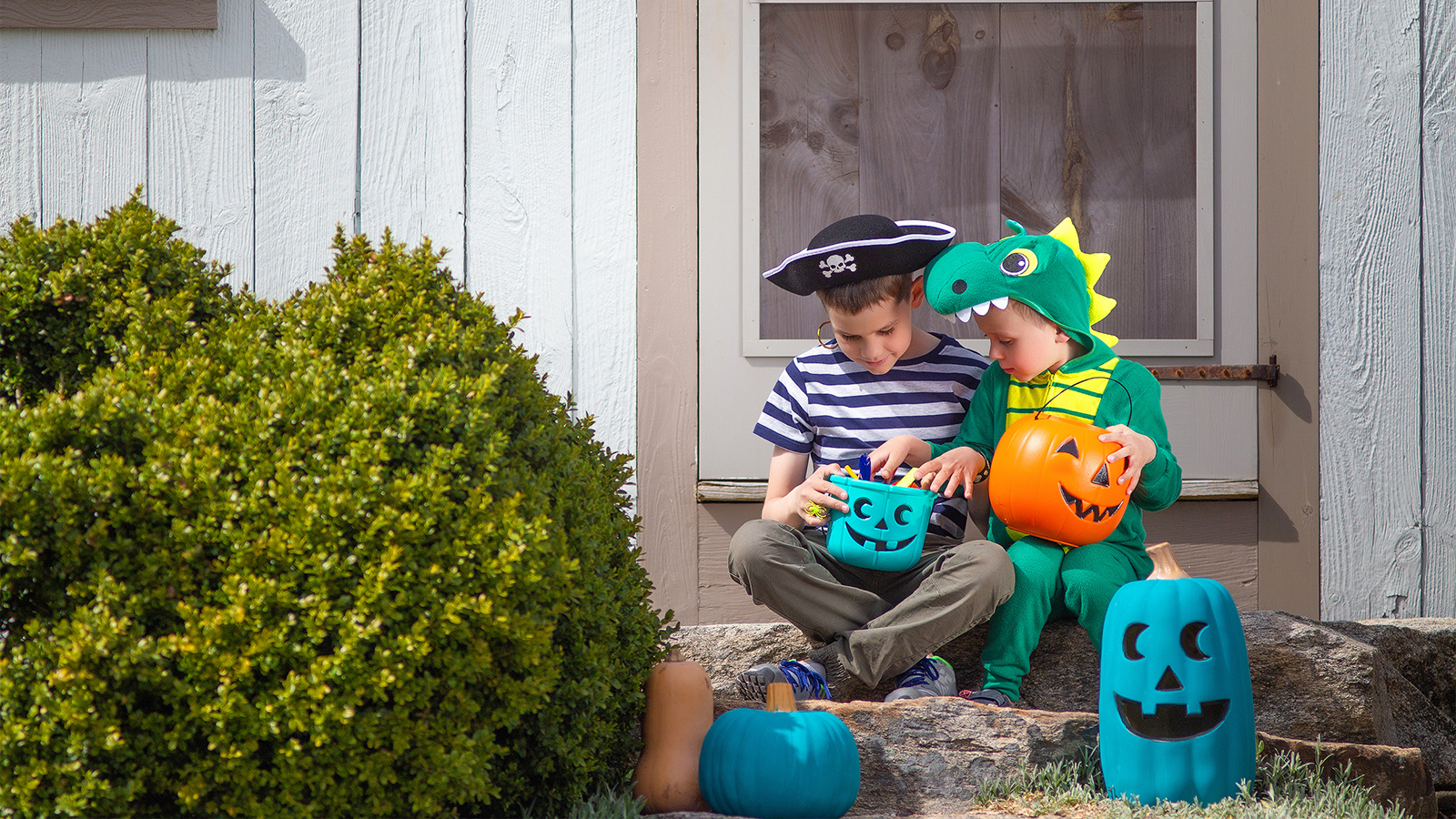 Two children sit on a porch, dressed in costumes, holding colorful pumpkin-shaped candy buckets, ready for Halloween.
