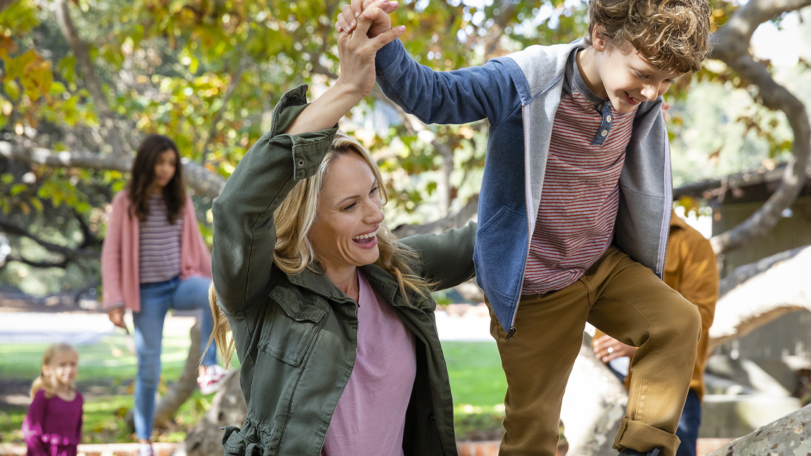 An adult helps a child climb a tree, both smiling and enjoying their time outdoors in a playful moment.