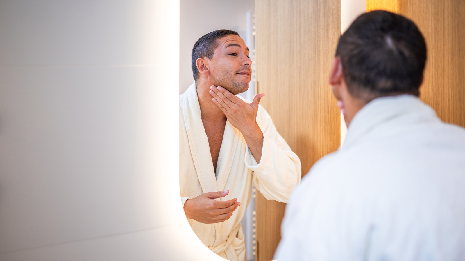 A man in a bathrobe examines his face in a mirror, reflecting personal grooming in a stylish bathroom.