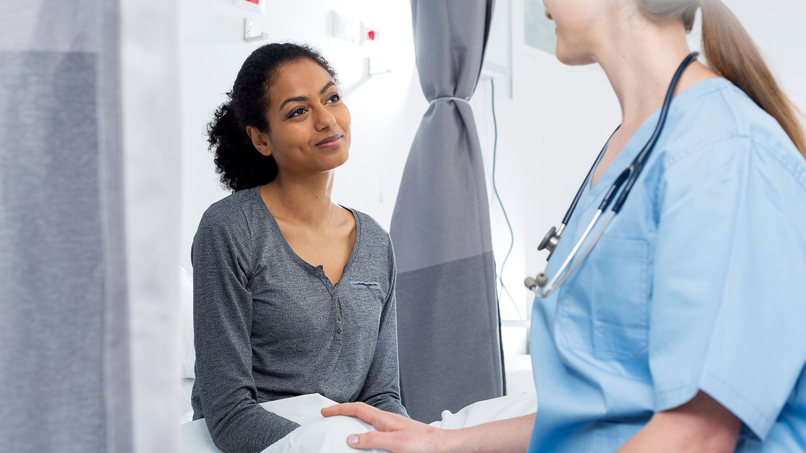 A nurse speaks with a patient sitting on a hospital bed, providing care in a clinical environment.