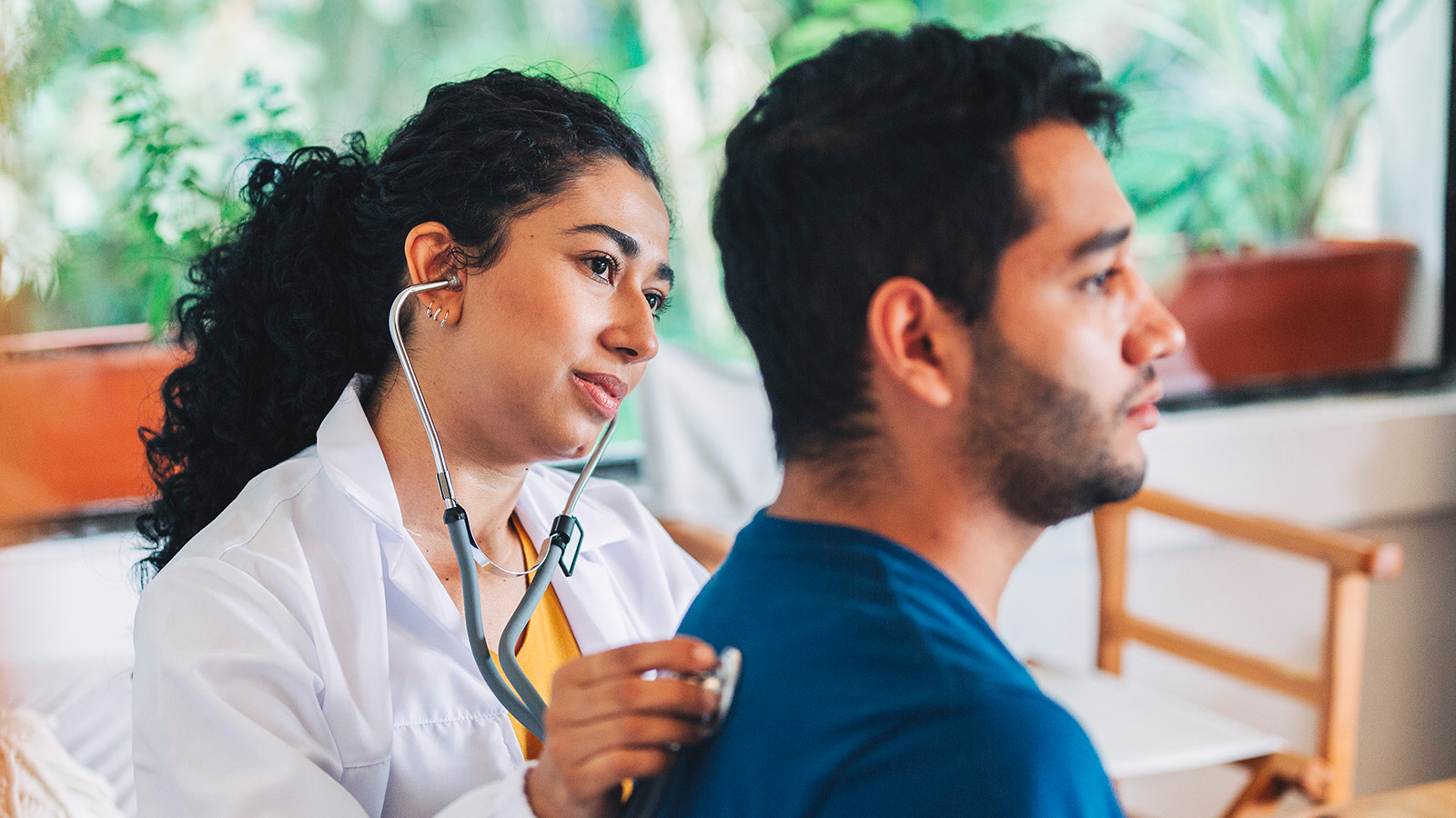 A healthcare professional listens to a patient’s back with a stethoscope in a bright, plant-filled room.
