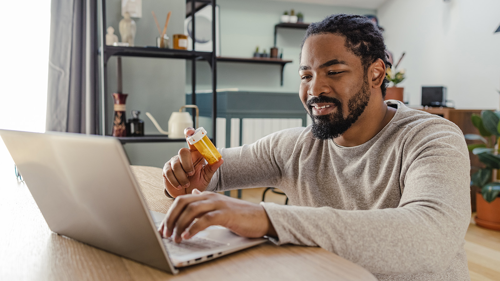 A man sits at a table, holding a prescription bottle while using a laptop, focused on health management.