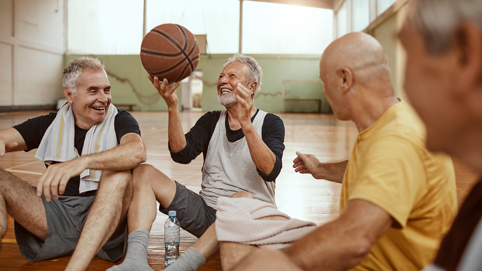 A group of older men relaxes on a basketball court, one preparing to throw a basketball while chatting.