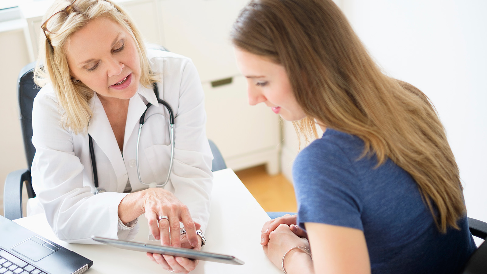 A doctor and patient engage in conversation, reviewing information on a tablet in a well-lit medical office setting.