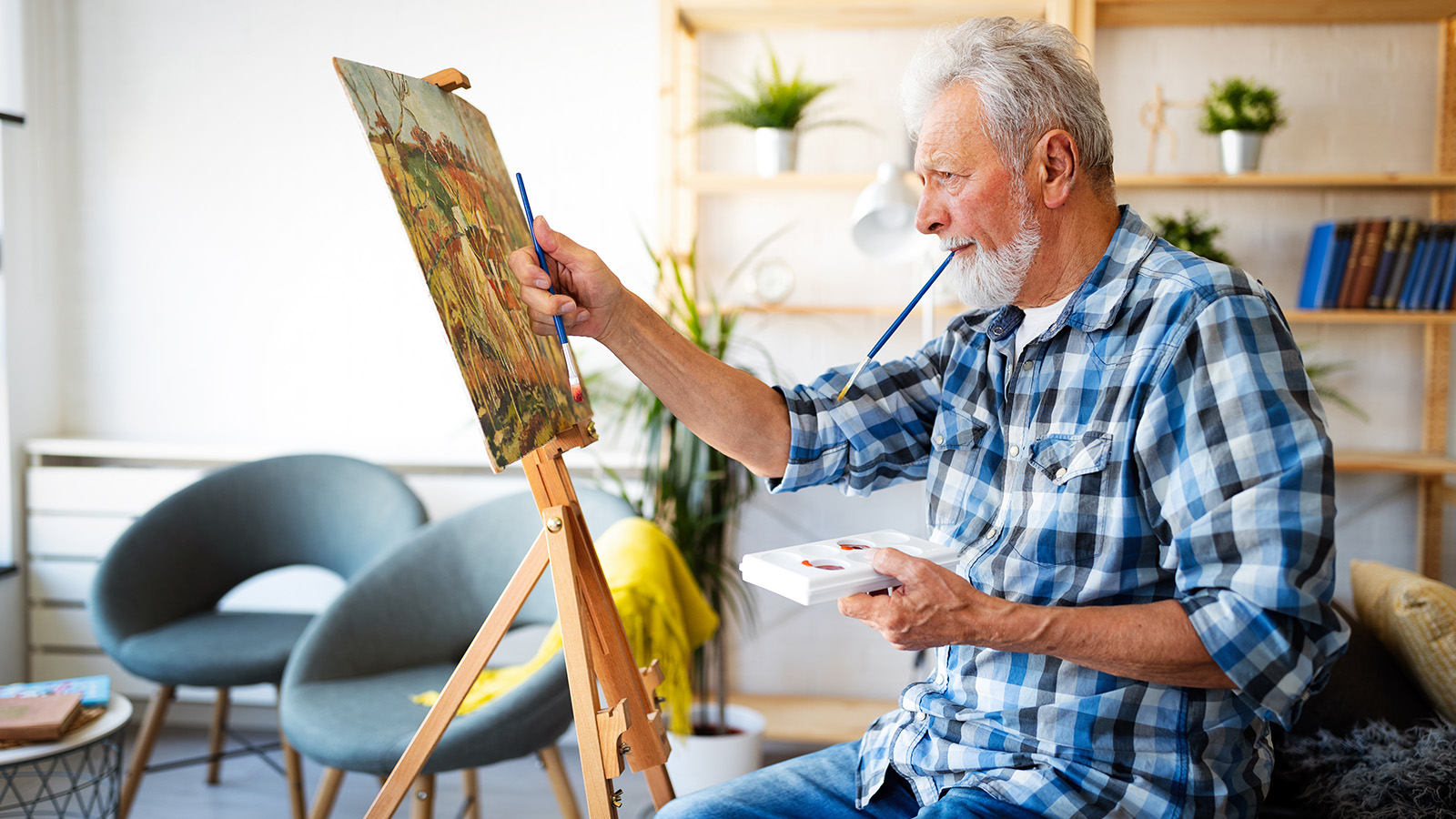 An elderly man paints on an easel, holding a palette with colors, surrounded by a cozy, well-lit room.