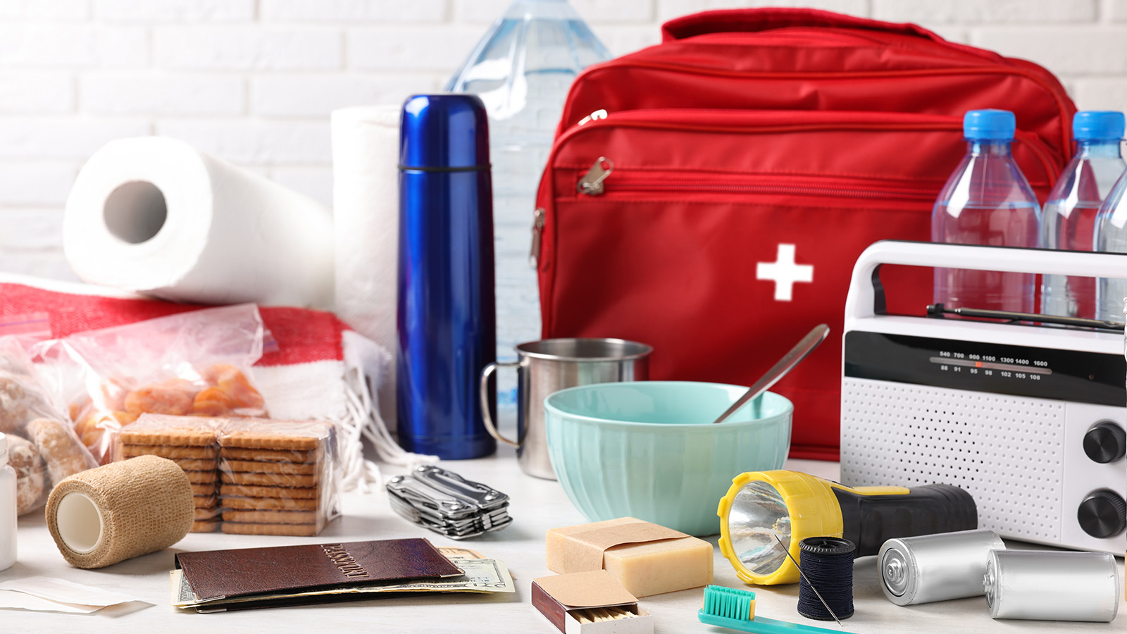 A red first aid kit surrounded by water bottles, snacks, a flashlight, and various emergency supplies on a table.