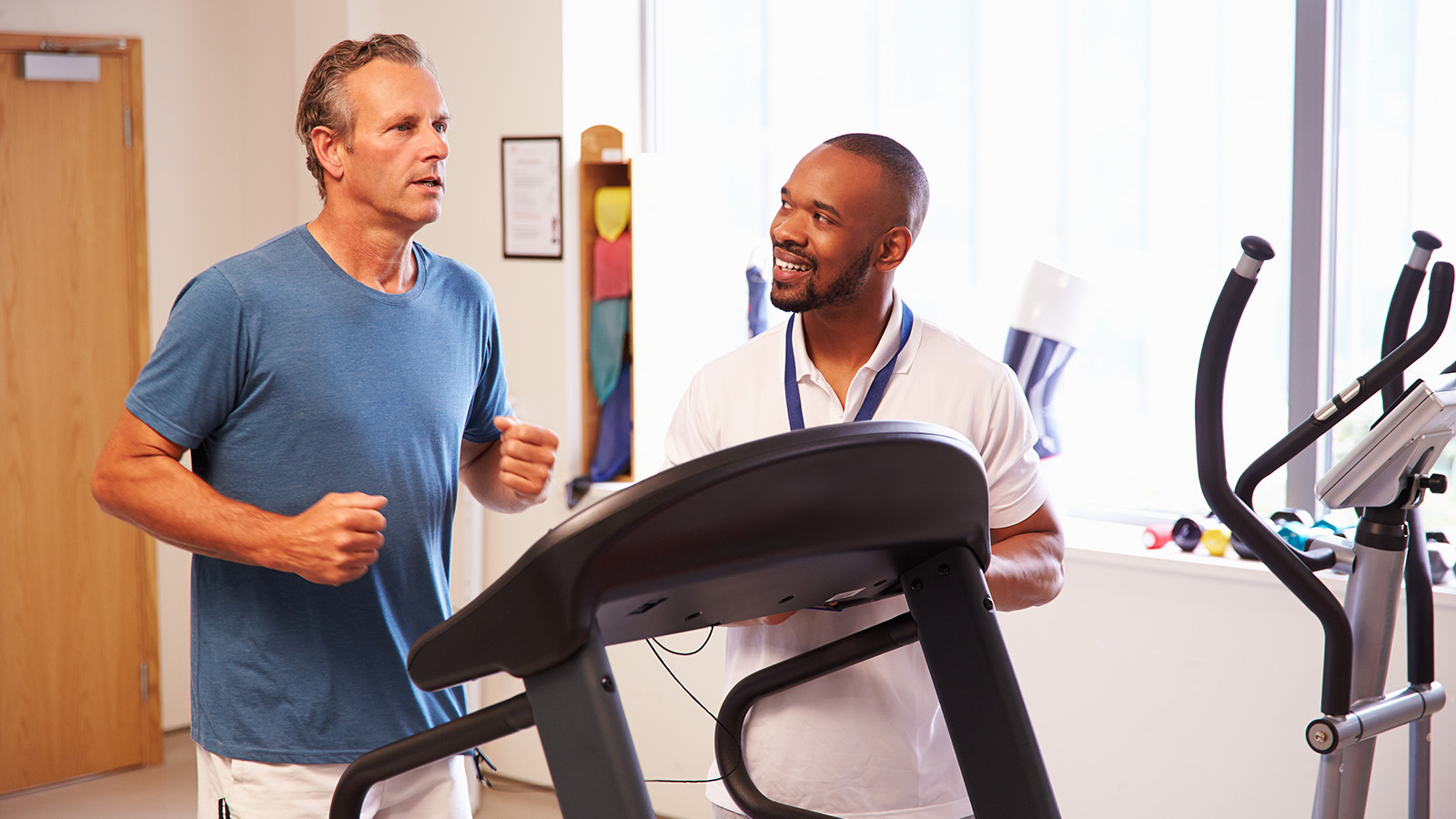 A man jogs on a treadmill while a trainer observes and provides guidance in a bright gym setting.