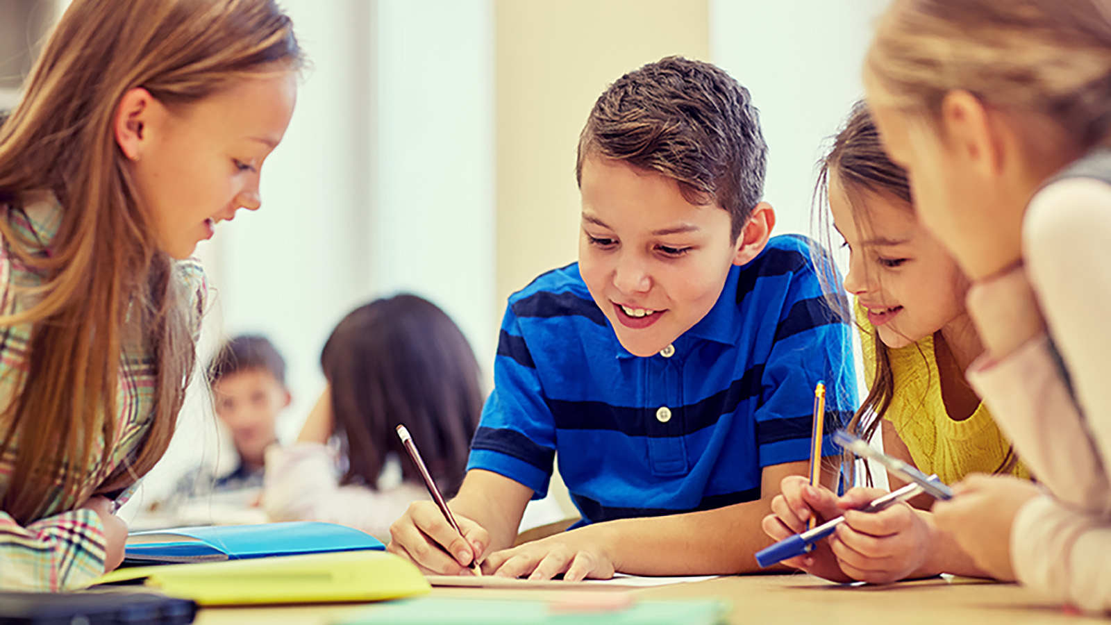 A group of children collaborates on a project at a table, with notebooks and pens scattered around them.