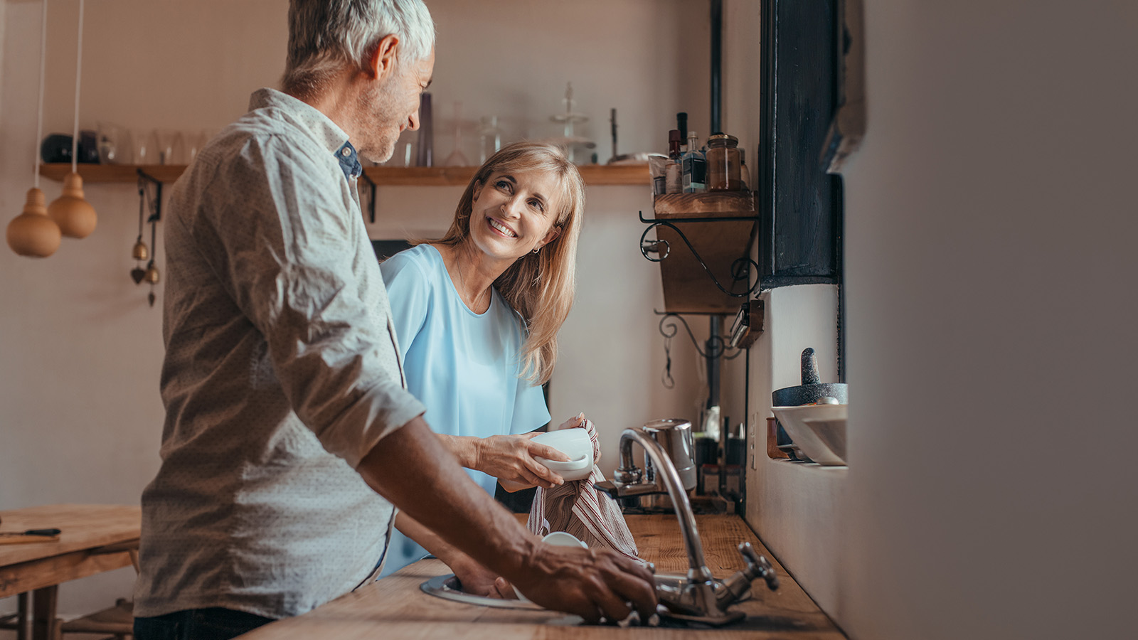 A man and woman are in a kitchen, with the woman holding a cup and the man washing dishes.