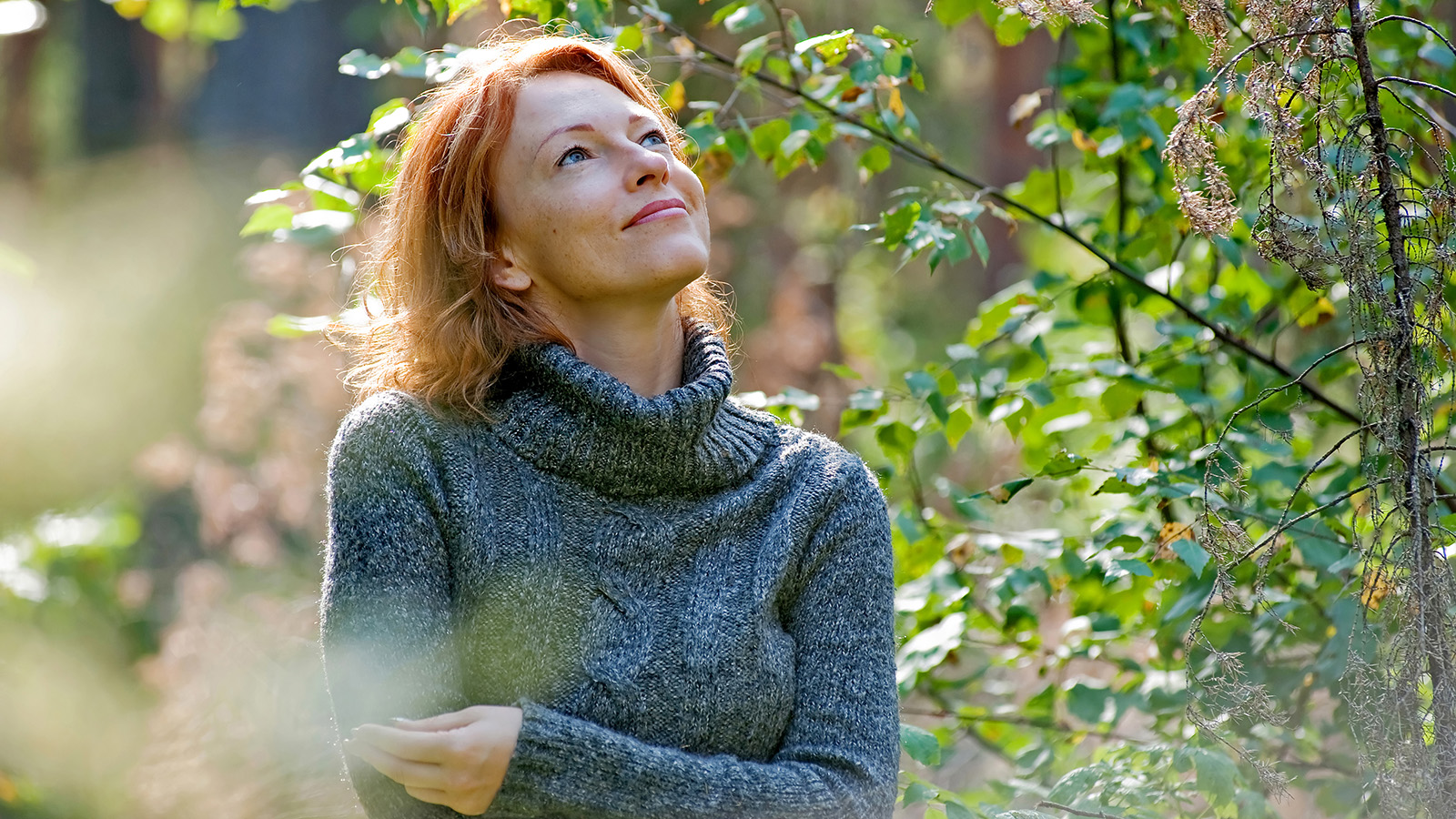A woman stands in a forest, looking contemplative, with greenery surrounding her in a natural setting.