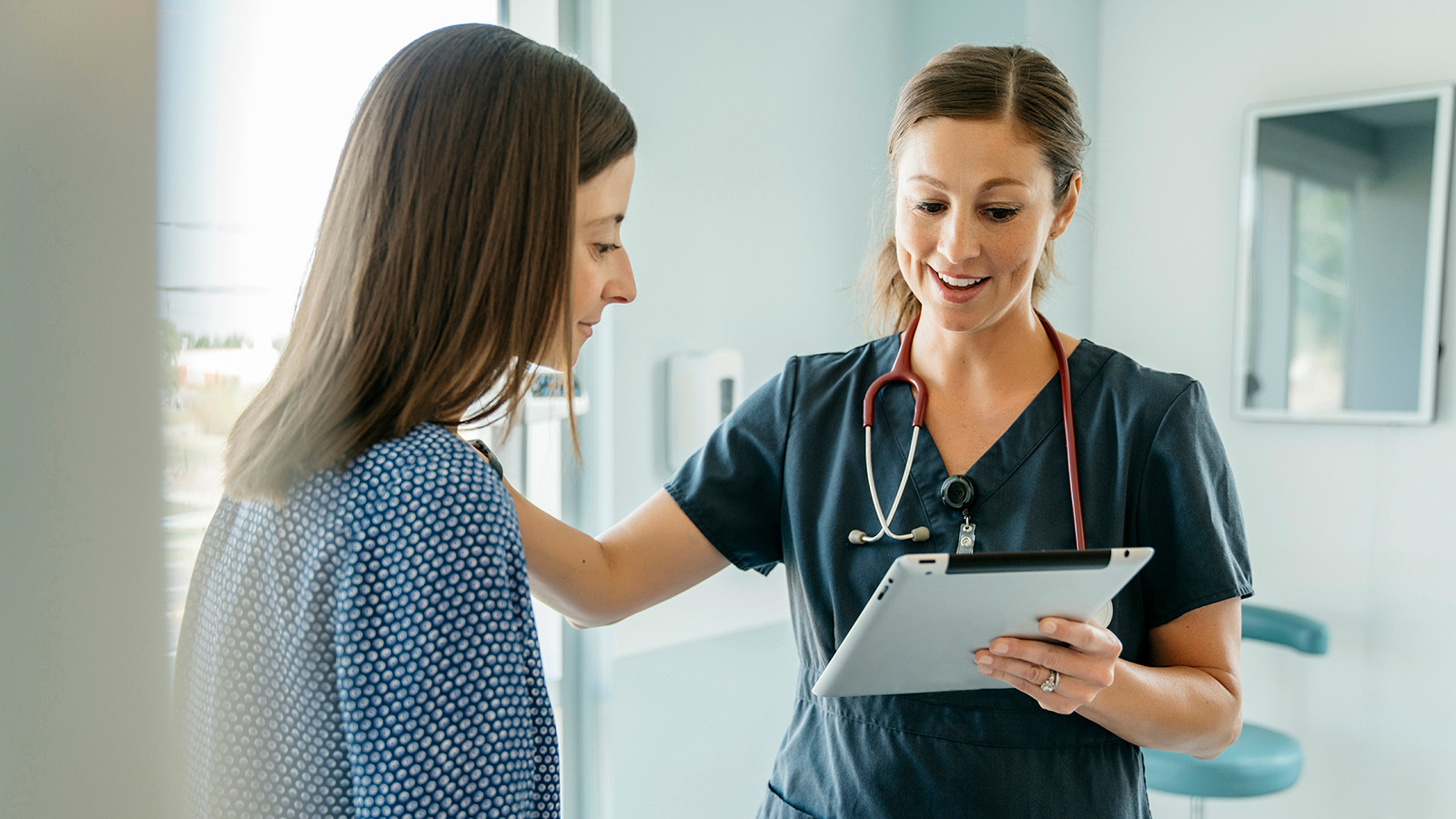 A woman is conversing with a healthcare professional, who is holding a tablet in a clinical environment.