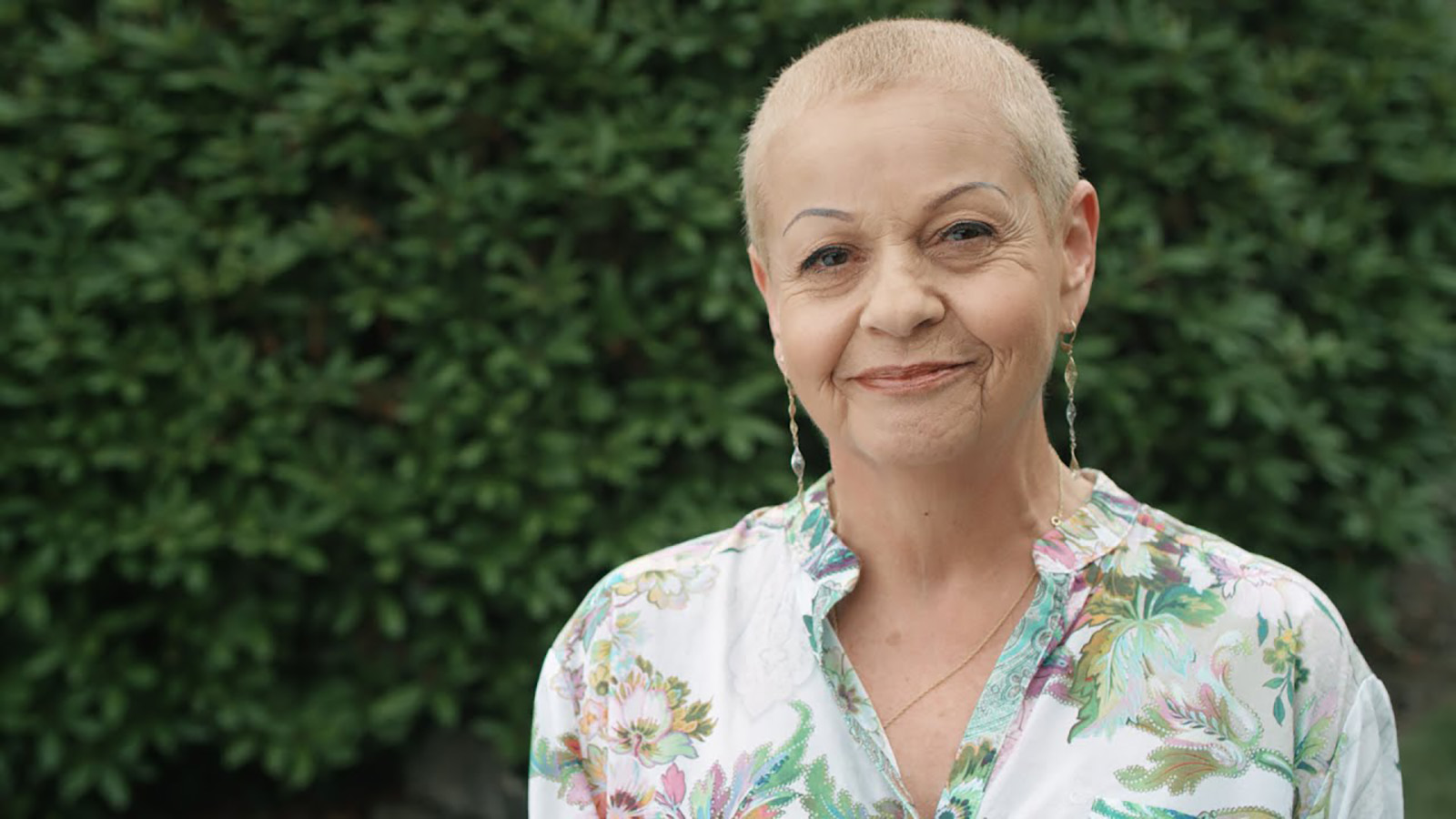 A woman stands in a forest, looking contemplative, with greenery surrounding her in a natural setting.