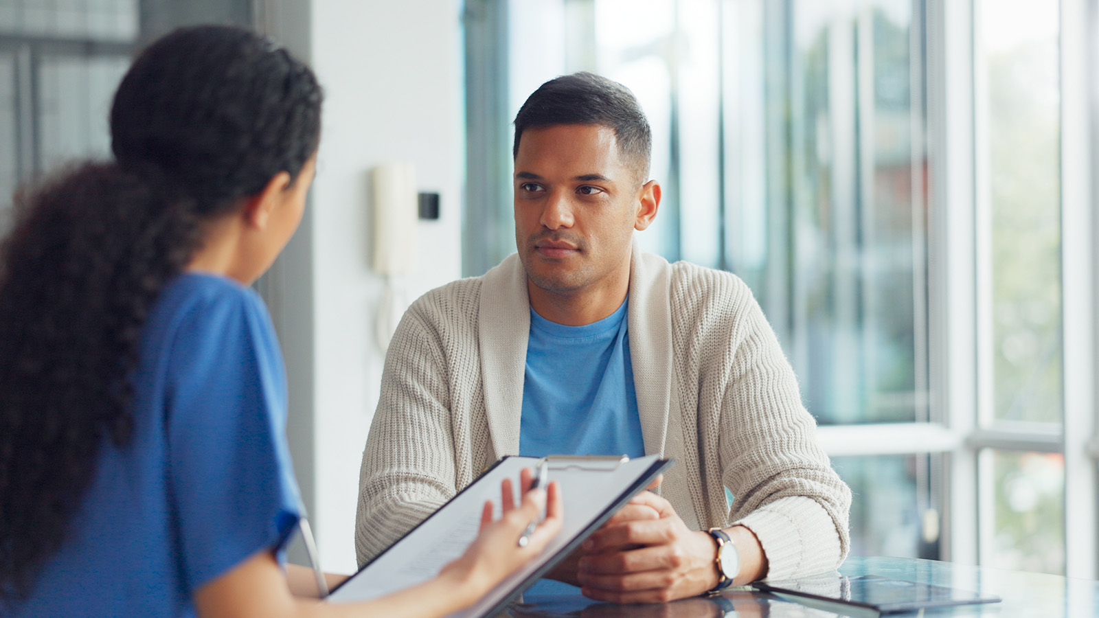 A woman in blue scrubs discusses paperwork with a man wearing a cardigan in a bright, modern office setting.