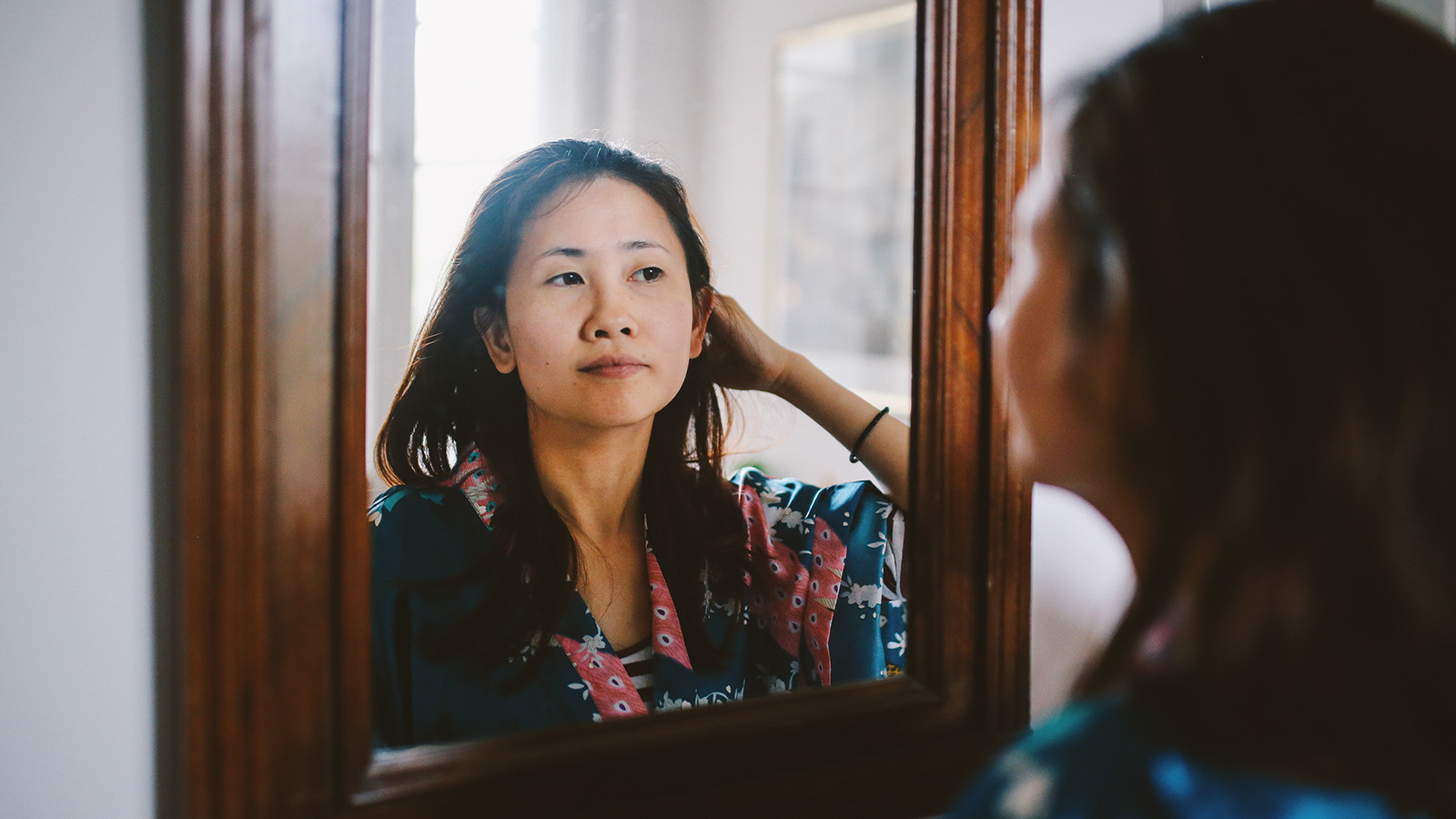 A woman in a colorful robe looks at her reflection in a mirror, adjusting her hair in a well-lit room.