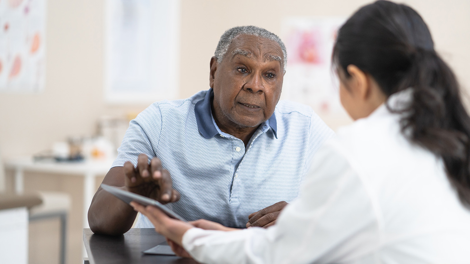 A healthcare professional in a white coat talks to an older man in a blue polo shirt at a medical office.
