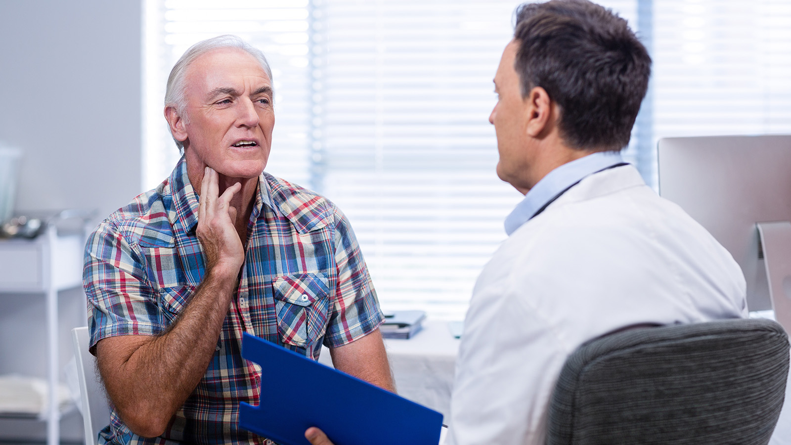 A man is engaged in conversation with a doctor, holding a blue folder, in a well-lit medical office.