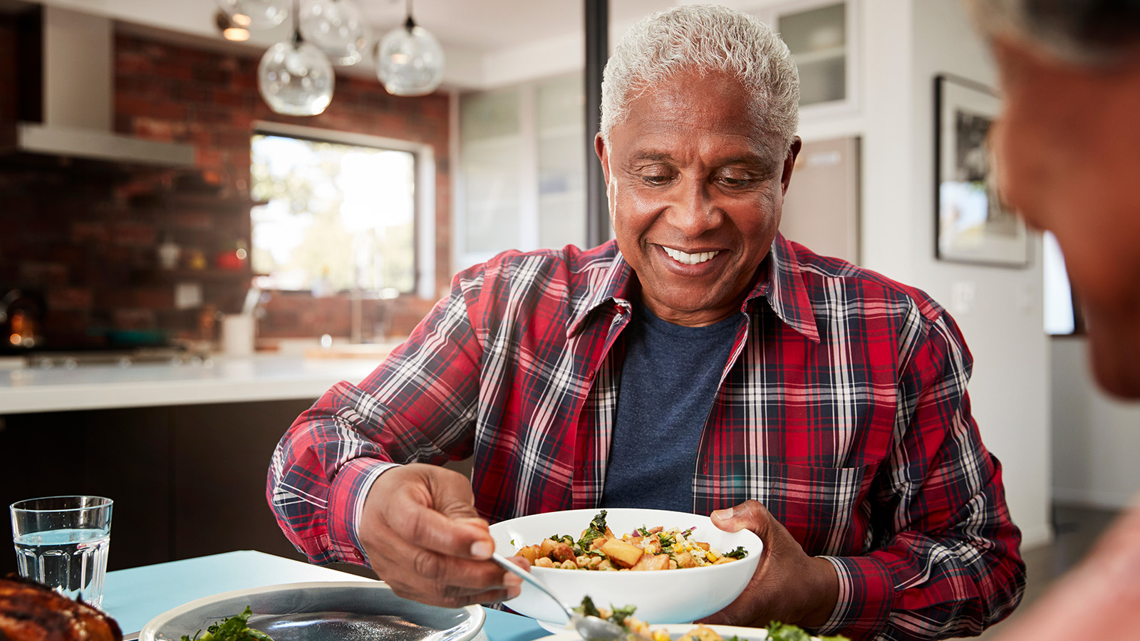 An older man holding a bowl of food at a dining table, with a woman seated nearby.