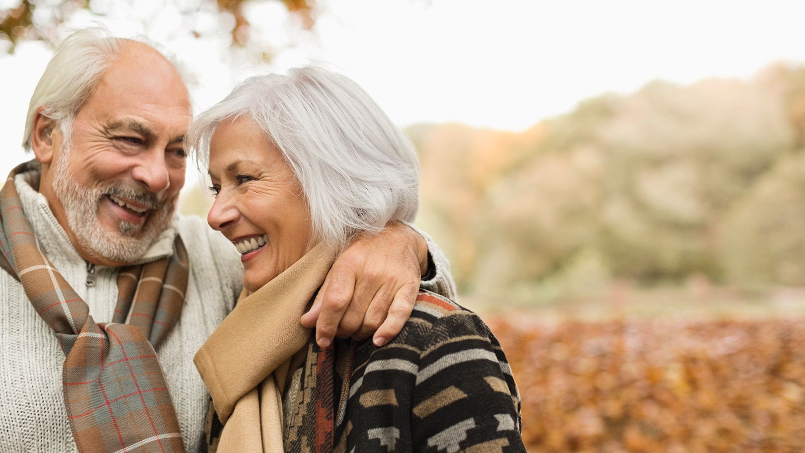 An elderly couple embracing outdoors, surrounded by autumn foliage, both wearing scarves and warm clothing.