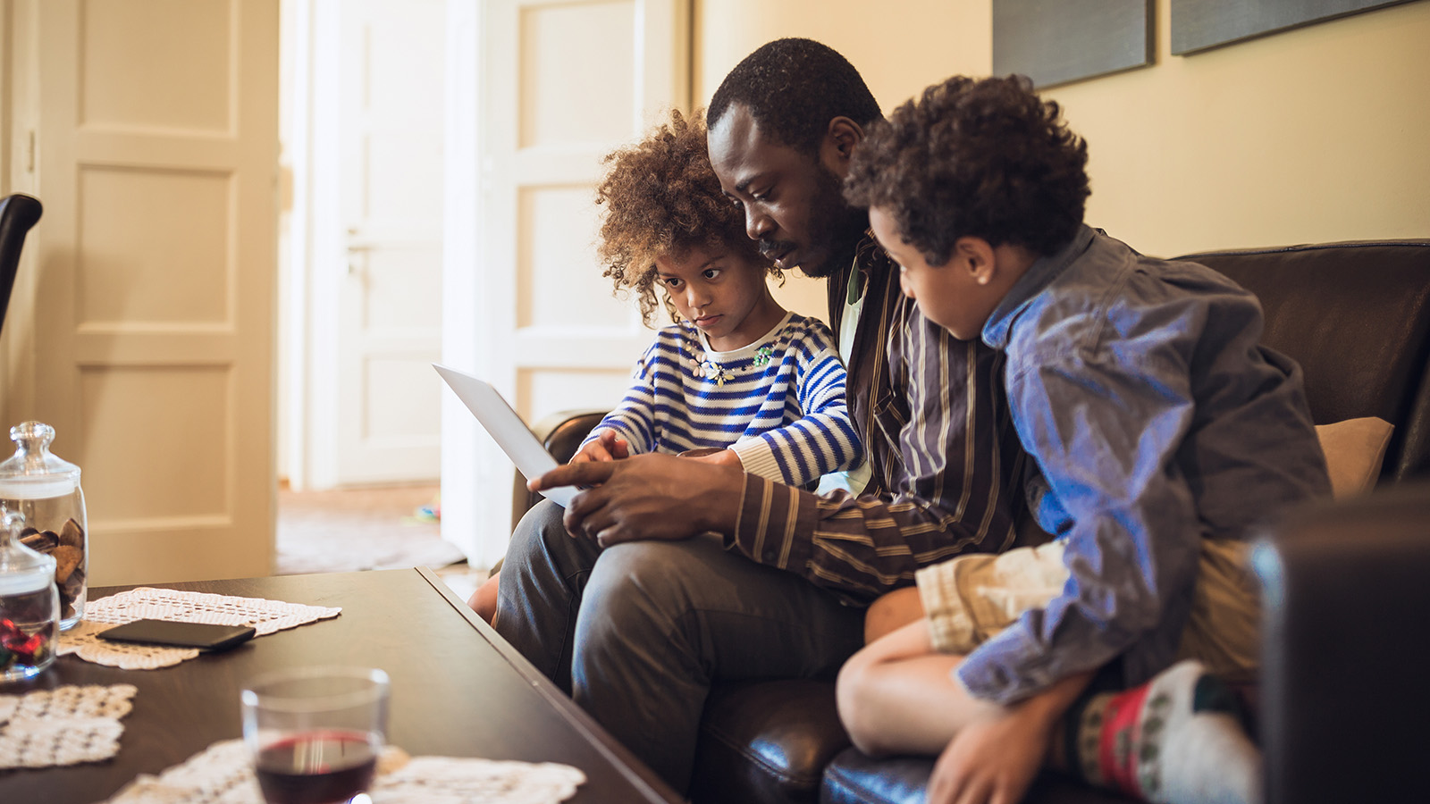 A parent sits on a couch with two children, reading a book together in a cozy living room.