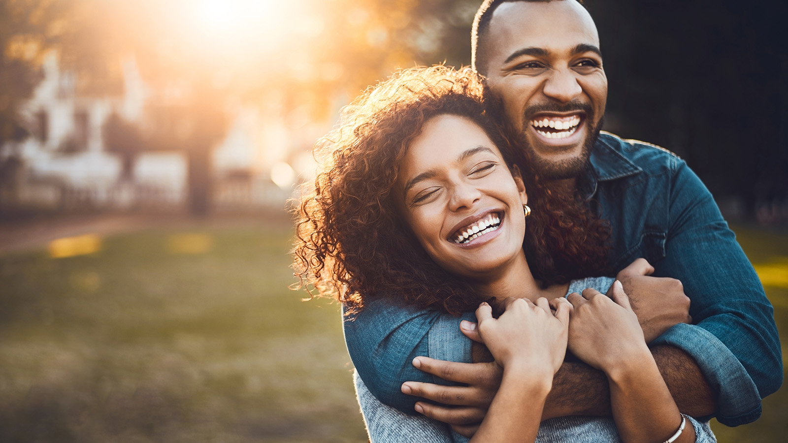 A couple embraces outdoors, surrounded by sunlight, showcasing their affection while standing close together in a park.